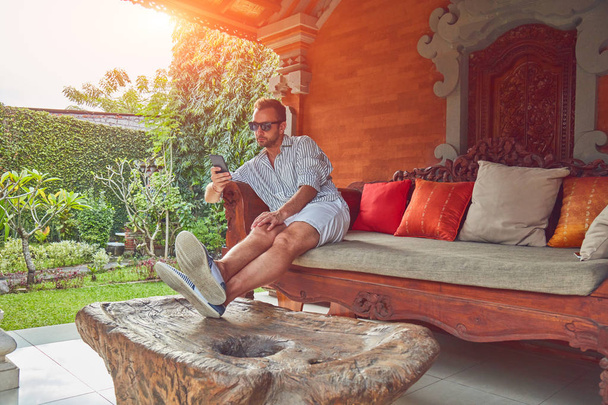 Homme utilisant un téléphone intelligent sur un canapé terrasse en été
. - Photo, image