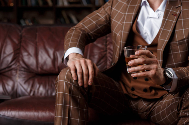 young successful man in a brown kosmtyum drinks whiskey. bearded businessman sitting in a library on a luxurious leather sofa and drinking cognac - Foto, Imagen