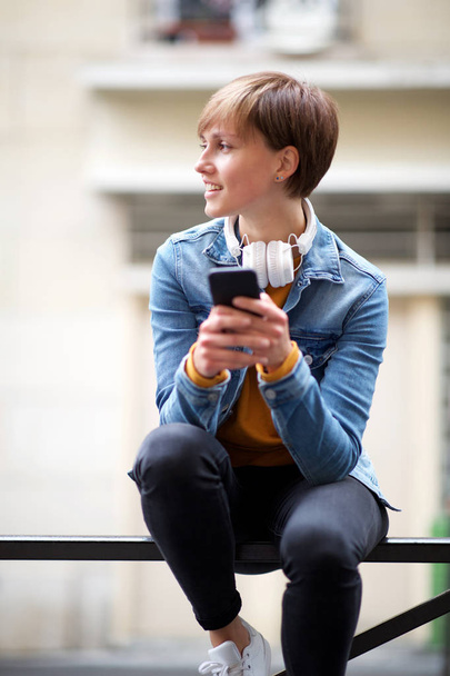 Πορτρέτο της δροσερό νεαρή γυναίκα που κάθεται με τα ακουστικά και έξυπνο τηλέφωνο - Φωτογραφία, εικόνα
