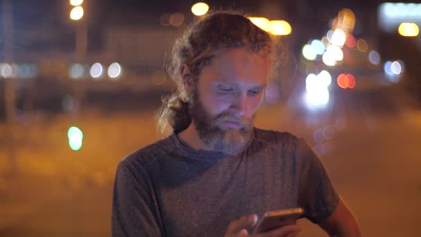 Close-up, portret van een knappe blanke bebaarde langharige jonge man die smartphone gebruikt op de achtergrond van het verplaatsen van Avondverlichting van een stad. - Video