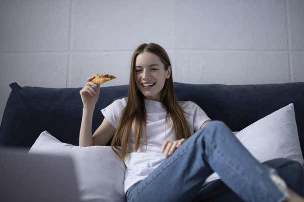 Happy young woman eating slice of hot pizza at home and watching - Foto, Imagen