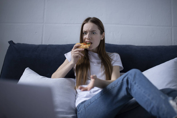 Happy young woman eating slice of hot pizza at home and watching - Фото, зображення