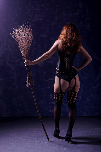 Sexy woman in a corset, stockings and with a broom posing on a dark background, rear view. Halloween concept. - Foto, imagen