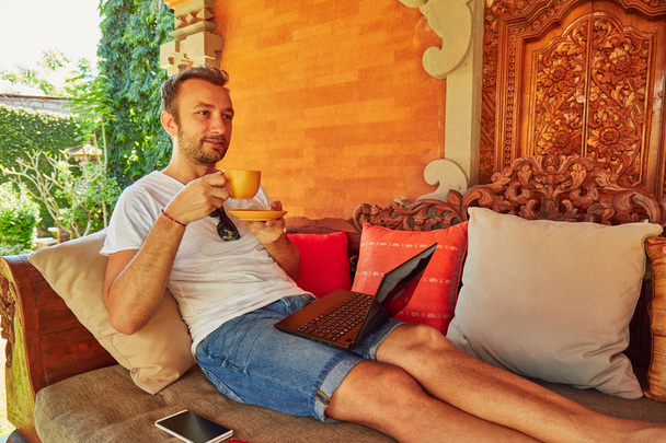 Man met koffie/thee en laptop op een veranda thuis. - Foto, afbeelding