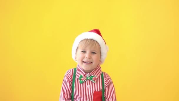 Happy child wearing Santa hat on yellow background. Funny kid showing thumbs up. Christmas winter holiday concept - Materiał filmowy, wideo