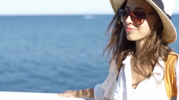 Hermosa viajera sonriente con sombrero de paja y gafas de sol relajándose en la playa, de pie contra un mar azul en un día soleado
 - Imágenes, Vídeo