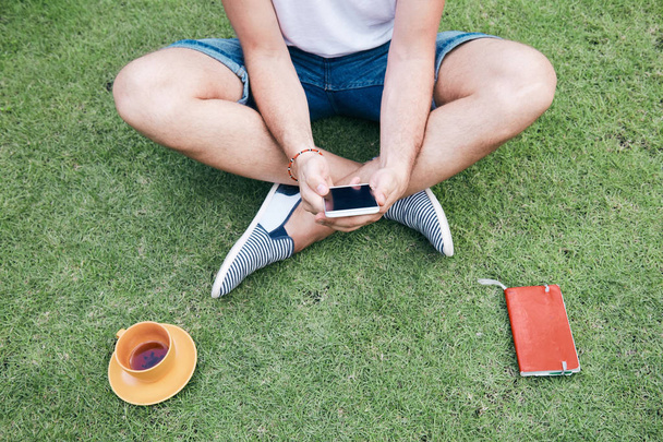 Man using smartphone and drinking coffee / tea on a grass. - Fotoğraf, Görsel