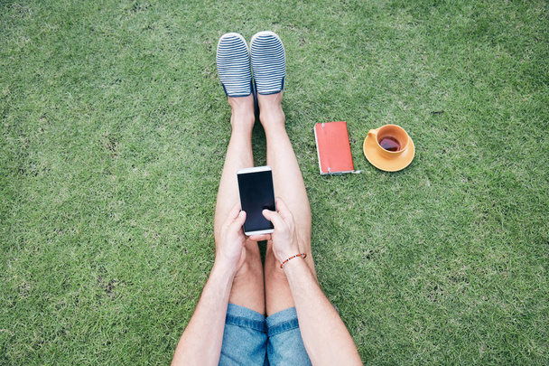 Man using smartphone and drinking coffee / tea on a grass. - Φωτογραφία, εικόνα