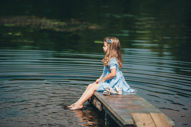 Little girl in blue dress sitting on a wooden pier near the water. Kid alone on the river. Side view - Φωτογραφία, εικόνα