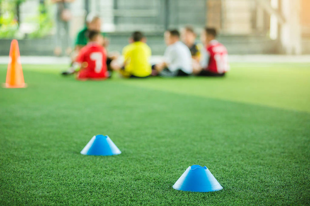 青いマーカーコーンに選択的な焦点は、ぼやけた子供の選手のトレーニングの背景を持つ緑の人工芝上のサッカートレーニング機器です。サッカーアカデミーのトレーニングクラスのための材料. - 写真・画像