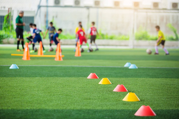 赤と黄色のマーカーコーンに選択的な焦点は、ぼやけた子供の選手のトレーニングの背景を持つ緑の人工芝上のサッカートレーニング機器です。サッカーアカデミーのトレーニングクラスのための材料. - 写真・画像