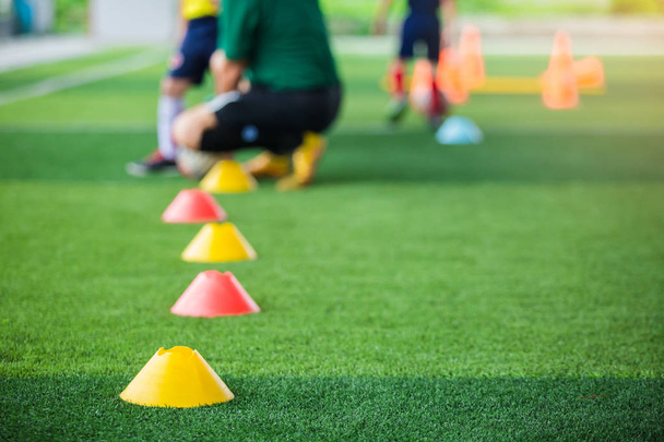 マーカーコーンへの選択的な焦点は、ぼやけたコーチと緑の人工芝上のサッカートレーニング機器は、子供の選手の背景を訓練しています。フットボールアカデミーのトレーニングクラスのための材料。垂直. - 写真・画像