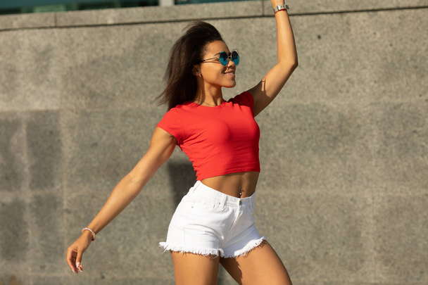 Fit mulatto woman posing wearing sunglasses, red crop top and white shorts on concrete wall background - Photo, Image