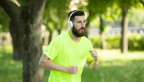 Young bearded man in headphones and sportswear training in park - Fotoğraf, Görsel