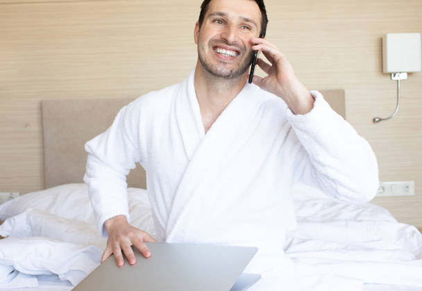 Young man in bathrobe using laptop and smartphone in bedroom at morning - Fotografie, Obrázek