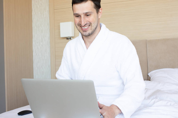 Young man in bathrobe using laptop in bedroom at morning - Photo, Image