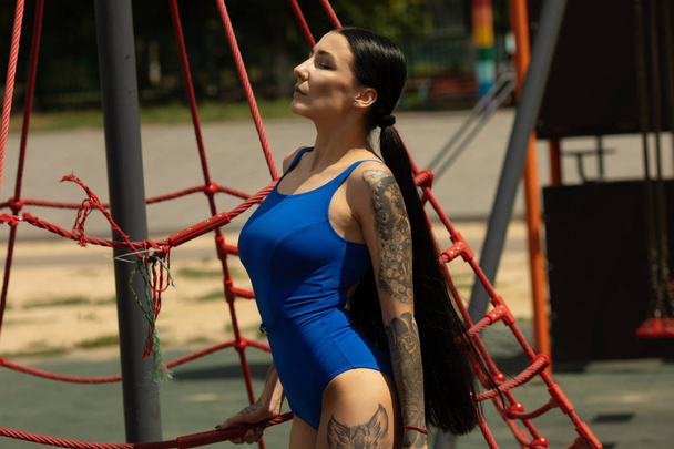 Fashion girl. Portrait of beautiful young tattooed hipster girl in blue swimsuit posing against urban background. - Zdjęcie, obraz