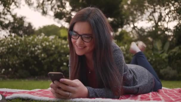 Portrait of relaxed smiling young woman lying on blanket in the park using smartphone - Video
