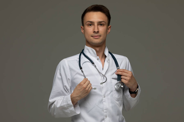 male doctor in white coat standing with stethoscope and looking at camera isolated on grey background. - Foto, immagini