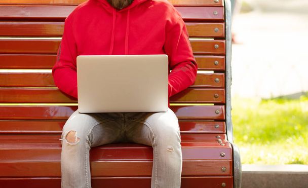 Young man in red jacket sitting on bench working with laptop in park - Zdjęcie, obraz