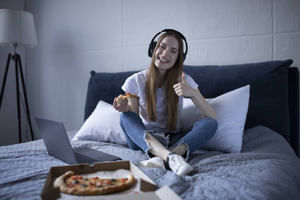 Χαρούμενη νεαρή γυναίκα τρώγοντας μια φέτα καυτή πίτσα στο σπίτι και βλέποντας - Φωτογραφία, εικόνα