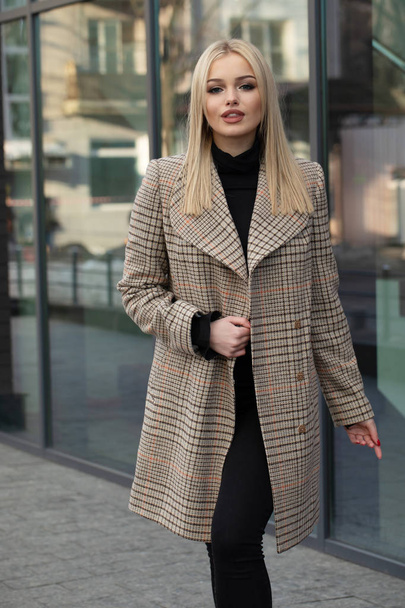 Blonde woman in warm beige woolen coat posing in city, modern building on background. - Fotografie, Obrázek