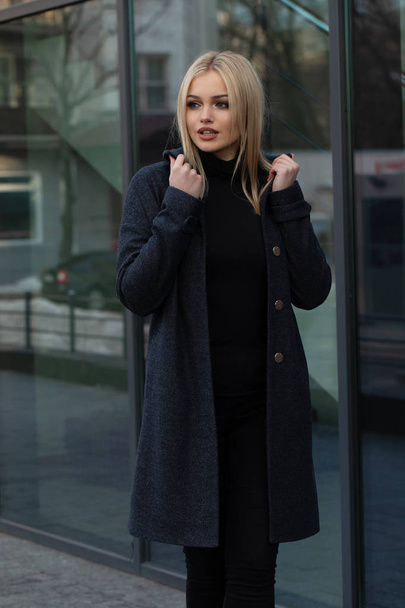 Portrait of young blonde woman in dark grey coat posing in city, modern building with festive shining lights on background. - Foto, Bild