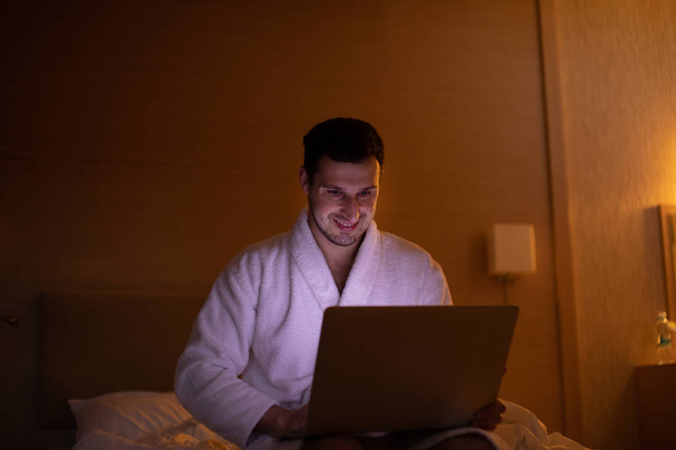 Young man in bathrobe using laptop in bedroom at night - Photo, image
