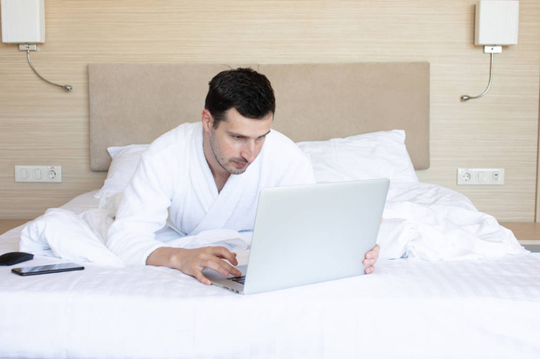 Young man in bathrobe using laptop in bedroom at morning - Zdjęcie, obraz