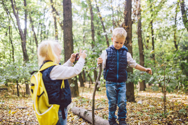 Children preschoolers Caucasian brother and sister take pictures of each other on mobile phone camera in forest park autumn. theme of hobby and active lifestyle for child. Profession photographer - 写真・画像
