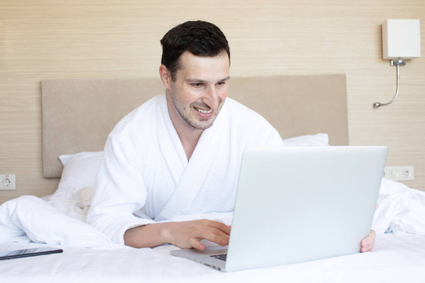 Young man in bathrobe using laptop in bedroom at morning - Zdjęcie, obraz