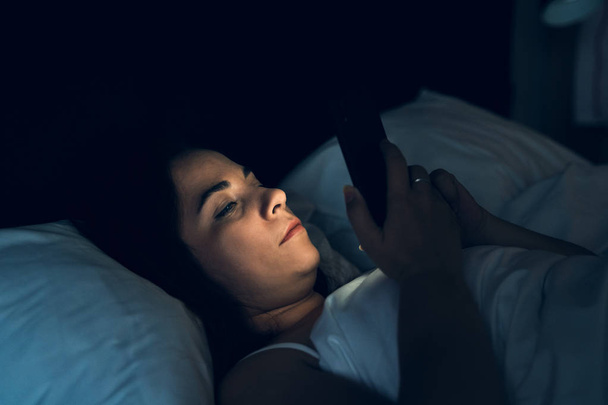 Uykusuzluk ve sosyal medya bağımlılığı kavramı. Genç kadın gece yatakta yatarken akıllı telefonu kullanıyor - Fotoğraf, Görsel