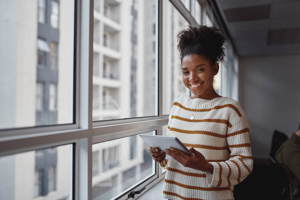 Retrato de uma jovem mulher de negócios afro-americana atraente com tablet digital em pé na frente das janelas do escritório
 - Foto, Imagem