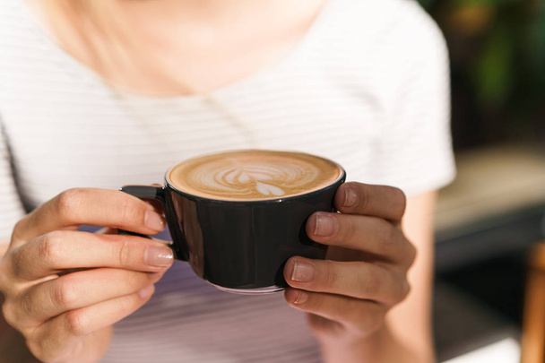 Περικομμένη εικόνα της καυκάσιας νεαρής γυναίκας κρατώντας φλιτζάνι καφέ λάτε στο καφέ - Φωτογραφία, εικόνα