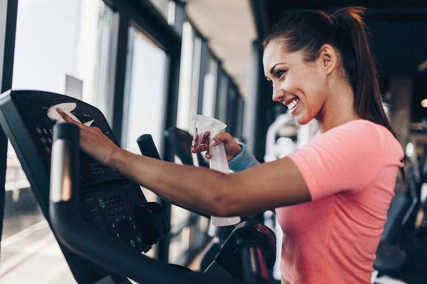 Young happy and smiling woman cleaning and weeping expensive fitness gym equipment with sprayer and cloth. - Photo, image