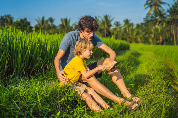 Dad and son sitting on the field holding tablet. Boy sitting on the grass on sunny day. Home schooling or playing a tablet - Foto, immagini