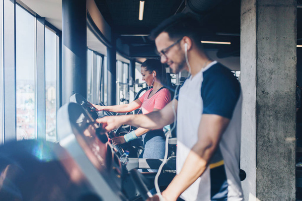 Young fit man and woman running on treadmill in modern fitness gym. - Foto, afbeelding