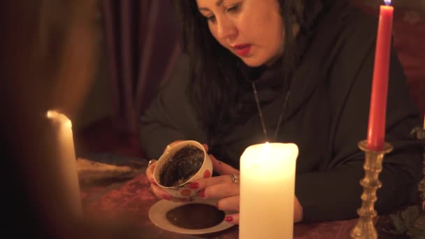 Fortune teller vrouw in donkere kamer met veel kaarsen geeft een uitleg van de toekomst en voorspelling van het lot aan een klant met koffie kopje dik sediment - Video