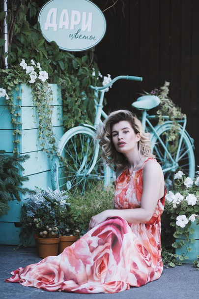 An attractive female in a floral pink dress posting near a bicycle and a flower gift sho - Foto, Imagen