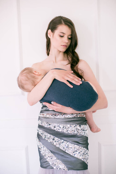 Young beautiful mother in evening dress on the floor holds the baby in her arms in a bright studio - Photo, Image