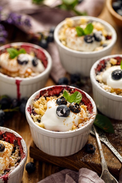 Blueberry crumble, stewed bilberries topped with crumble of flour, butter and sugar  with addition natural yogurt in a baking dishes on a wooden table, close-up. Homemade fruit dessert - Photo, Image