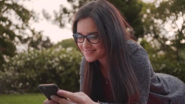 Portrait of an attractive smiling young woman lying in park texting messages on smartphone - Imágenes, Vídeo