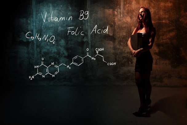 Sexy meisje of secretaresse of vrouwelijke student presenteren hand getekende chemische formule van vitamine B9 - Foto, afbeelding