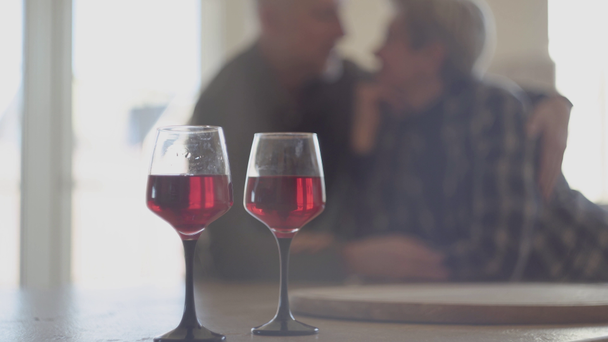 Twee glazen rode wijn en op de achtergrond is silhouet van een ouder stel. man vasthouden met liefde vrouw en kus haar neus. - Video