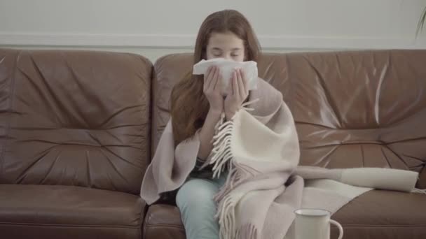 Small sick girl blowing her nose in the sofa, tablets, pills and cup on the table. - Materiał filmowy, wideo