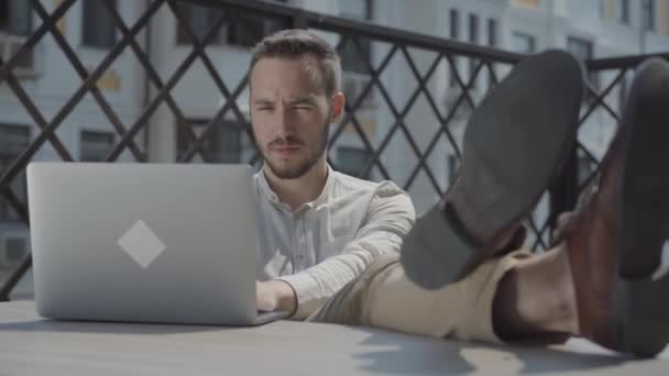 Bearded young man sitting at the table on the terrace with his feet on the table, the laptop lying near. Concept of freelance, distant work. The guy resting outdoors, video  - 映像、動画