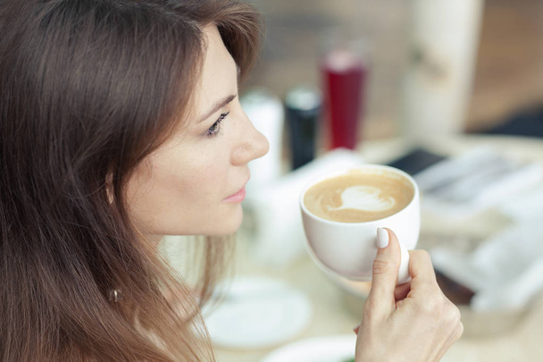 close portrait of a beautiful young brunette woman with a cup of cappuccino at the table - Photo, image