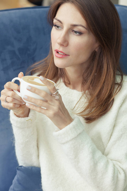 Lähikuva muotokuva kaunis nuori nainen viihtyisä pehmeä valkoinen pusero. Tyttö istuu nojatuolissa ja pitää kahvimukia käsissään.
. - Valokuva, kuva