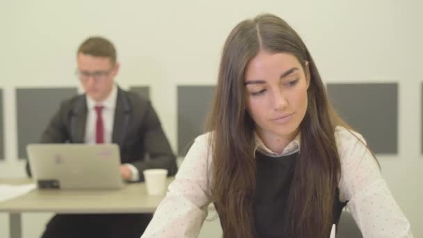 video of thoughtful young woman in formal wear sitting in foreground in office while her male colleague in suit working with laptop on the background - Video, Çekim