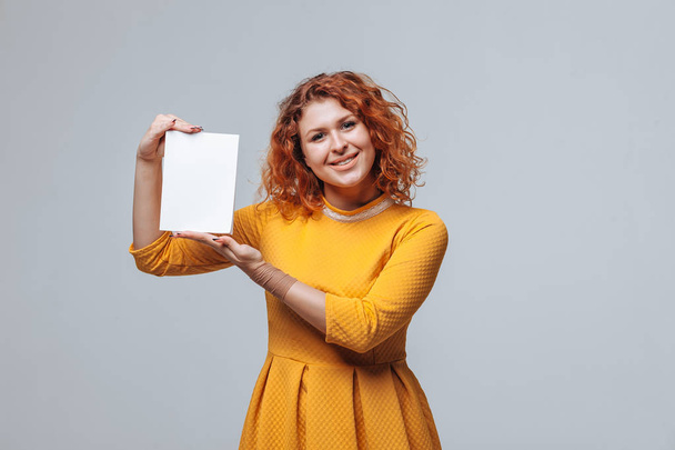 Red-haired girl holding a white book on a light gray background. - Zdjęcie, obraz